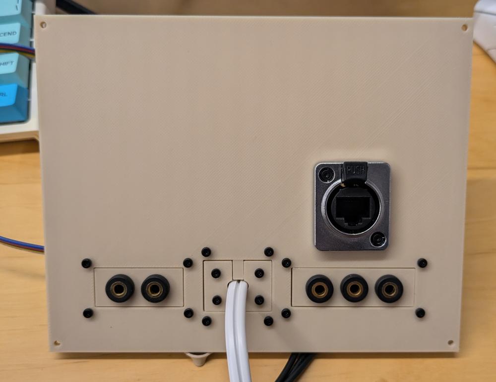 A flat 3d printed panel with screw holes in it's 4 corners. It has a group of 3 3.5mm audio jacks on the right, a second group of 2 audio jacks on the left, and a power cord coming out the middle. Above there is an ethernet port