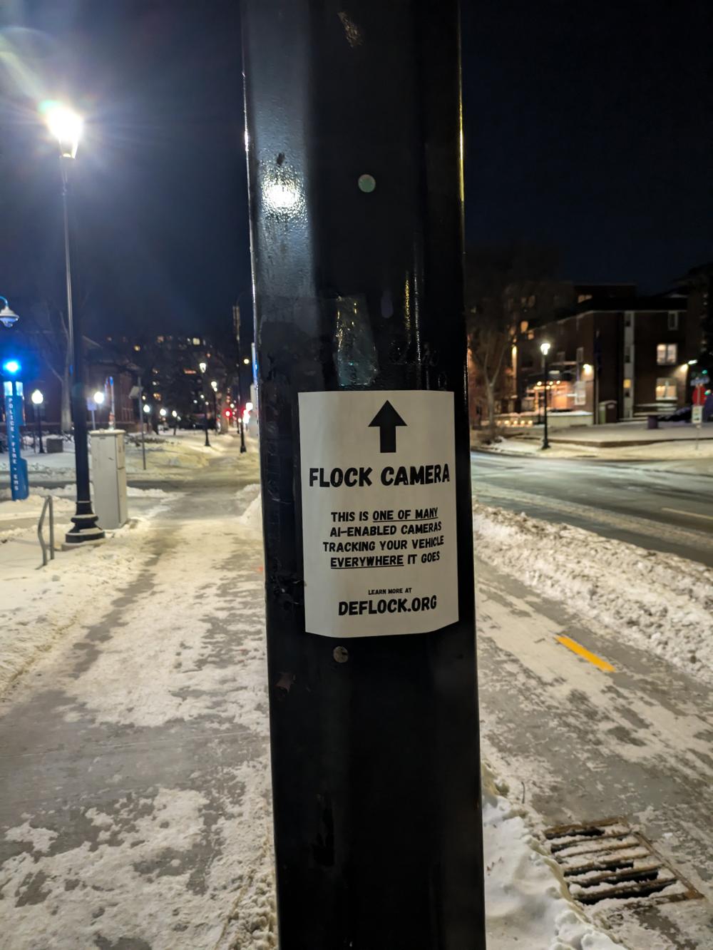 Sign on a pole with an arrow pointing. The sign reads "Flock camera. This is one of many AI enabled cameras tracking your vehicle everywhere it goes. Learn more at deflock.org